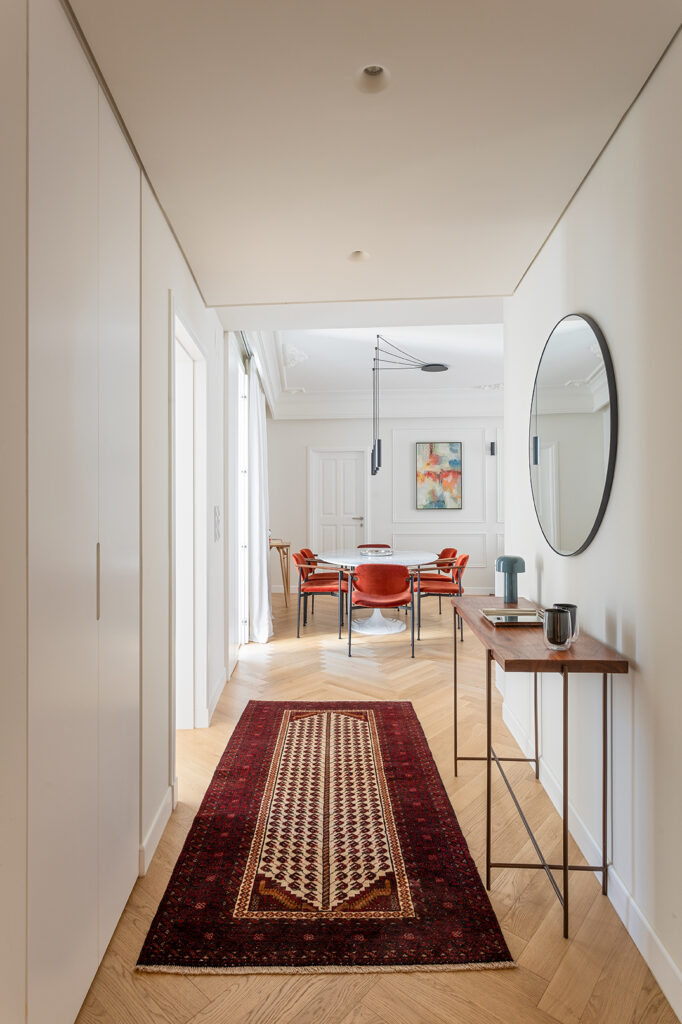Entrance hallway with herringbone wooden floor, white built-in cupboards, wooden console with round mirror and red carpet runner overlooking the dining room in Lisbon.