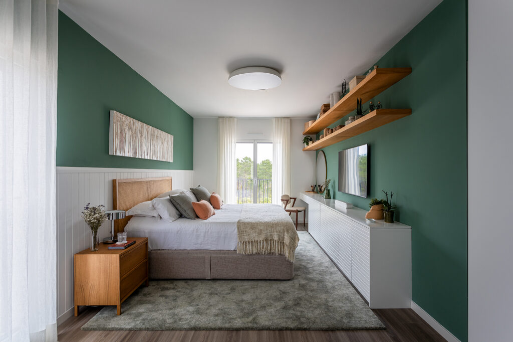 Decorating a double bedroom with green-painted walls, a straw headboard, white wainscoting, wooden bedside tables and a hanging TV cabinet with linear shelves in an apartment in Lisbon.