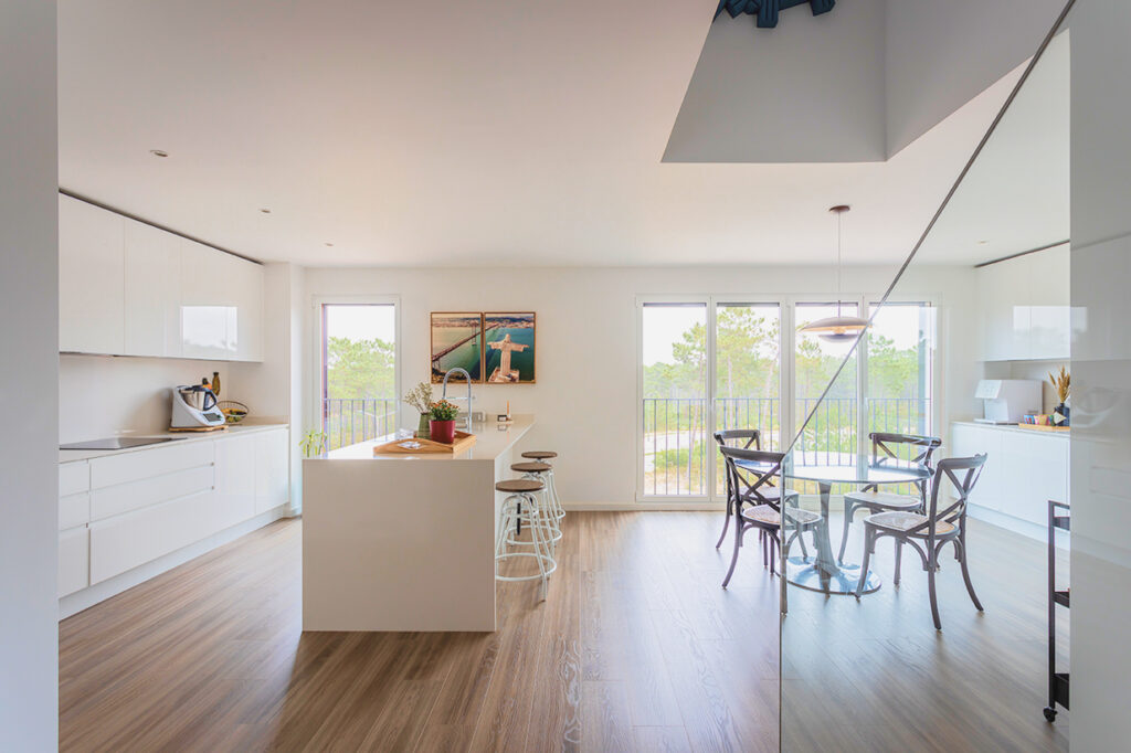 Refurbished kitchen in white tones with central island, high stools and integrated round glass dining table, overlooking nature in a project by CW Arquitetura in Lisbon.