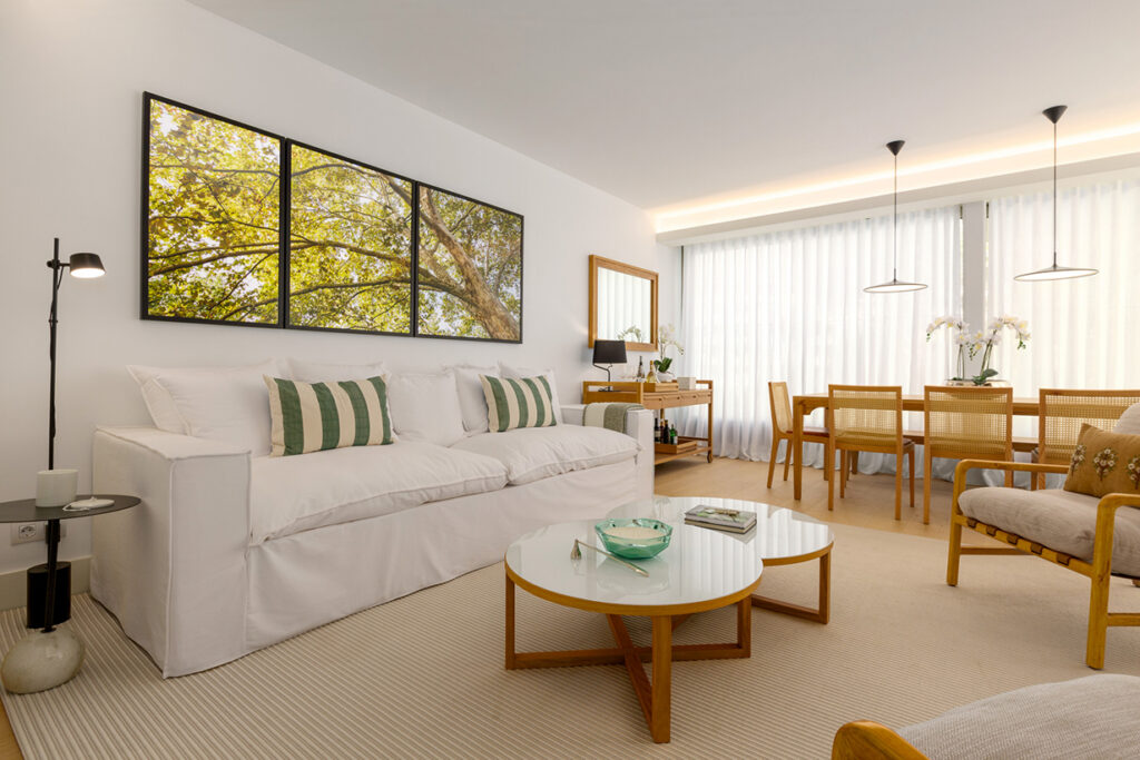 Detail of a living room decoration with a white sofa, green striped cushions, round coffee tables and a triptic of canvases with photographs of trees on the wall.