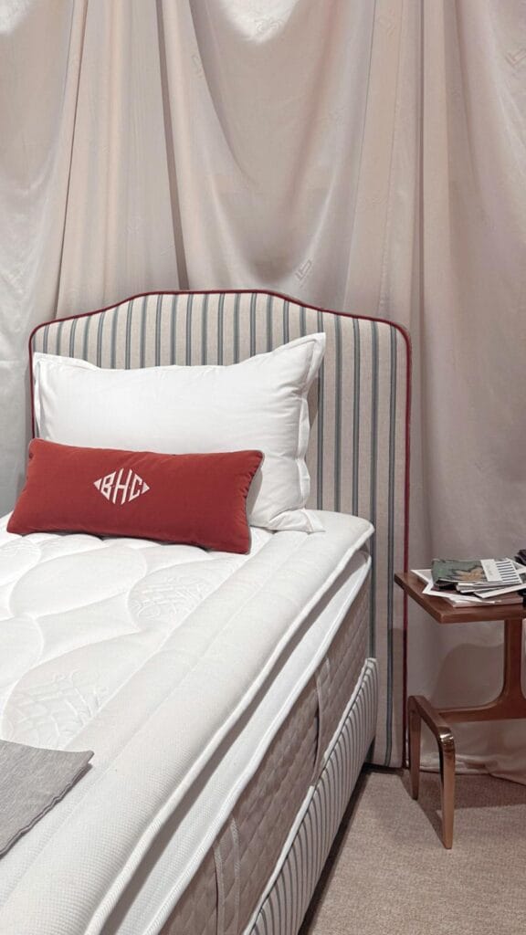 Cama de casal com cabeceira estofada listrada, roupa de cama branca e almofada bordada em tom terracota.