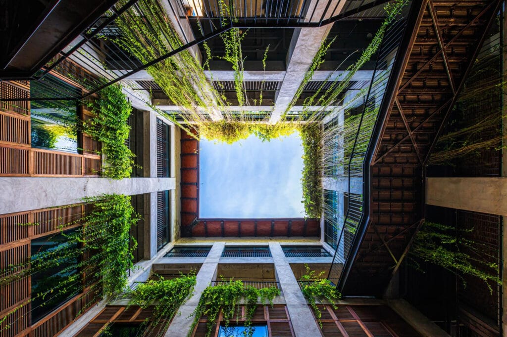 Atrio central de edificio com design biofilico, vegetacao suspensa, madeira e abertura para o ceu.