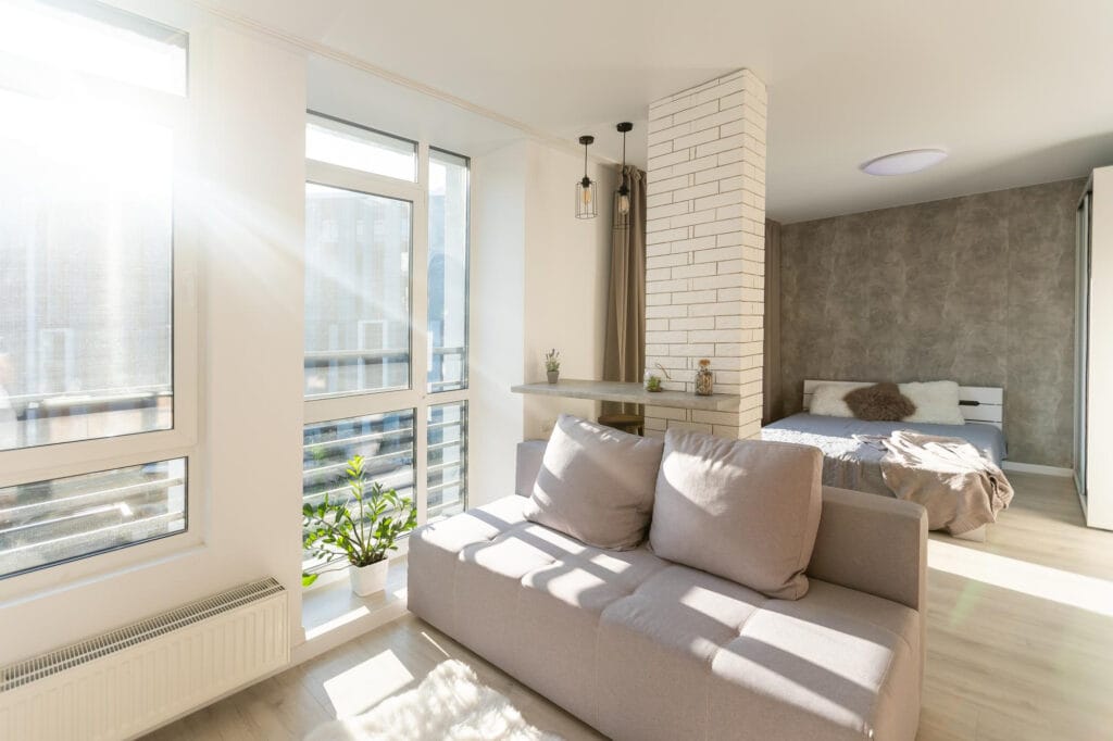 Sala de estar integrada com quarto recebendo abundante entrada de luz natural pelas grandes janelas em apartamento compacto em Lisboa.